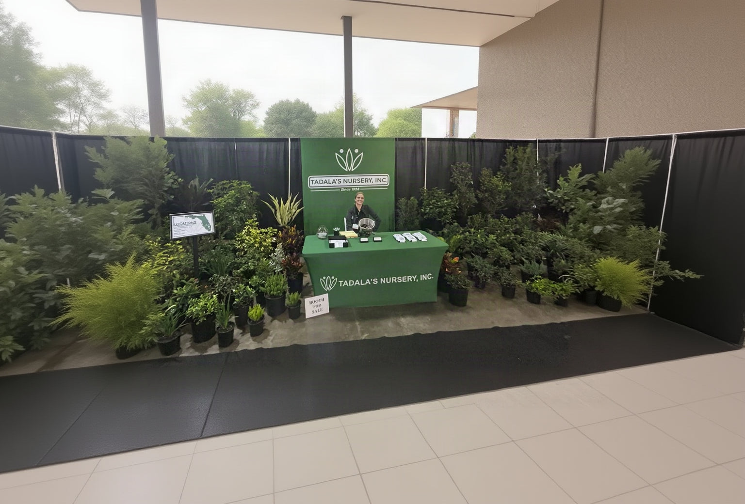 A booth display for Tadal's Nursery, Inc. featuring a green branded table and backdrop with a person standing behind it, surrounded by many potted plants at what appears to be a trade show or exhibition.