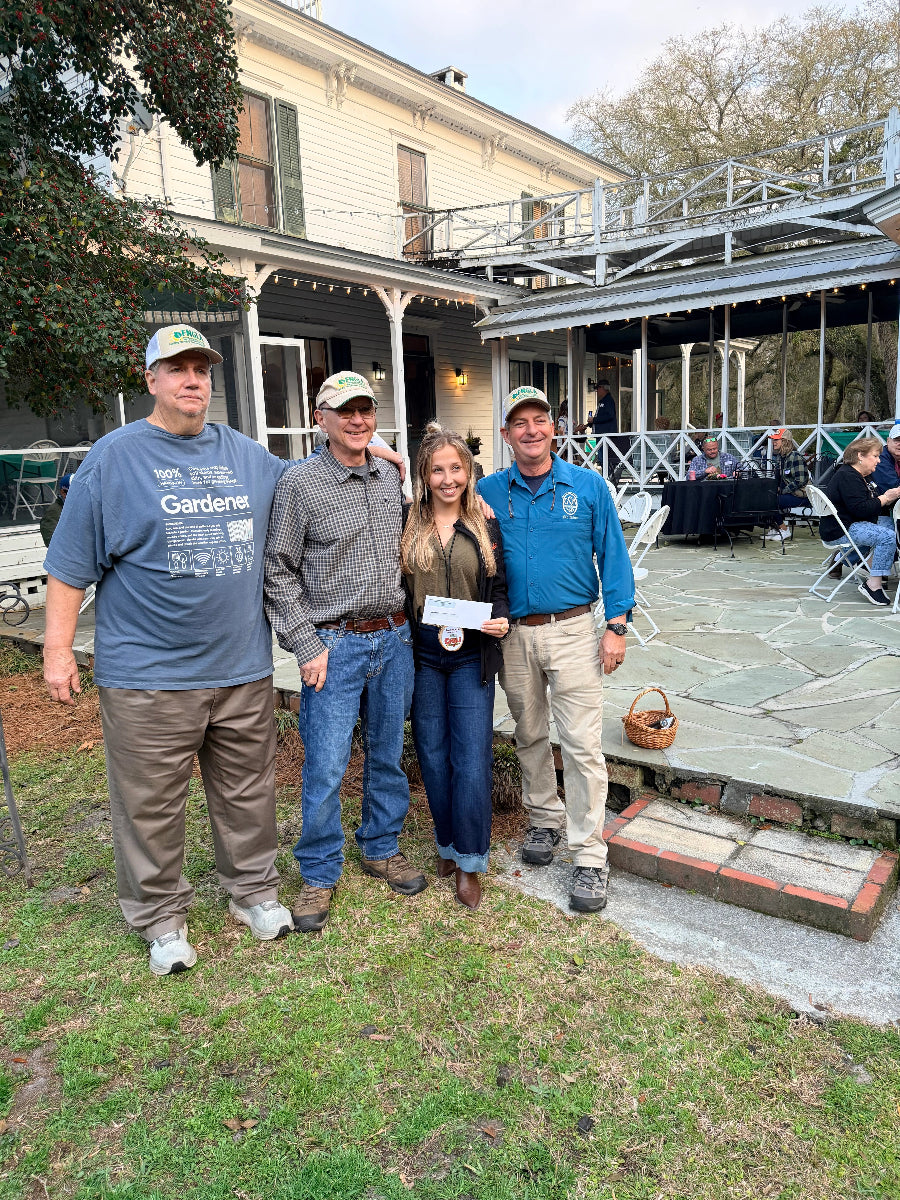 Four people standing together outdoors in front of a house and patio, holding a white certificate. Two men wear blue t-shirts, one man wears a gray patterned button-up shirt, and the woman wears a brown jacket and blue jeans.