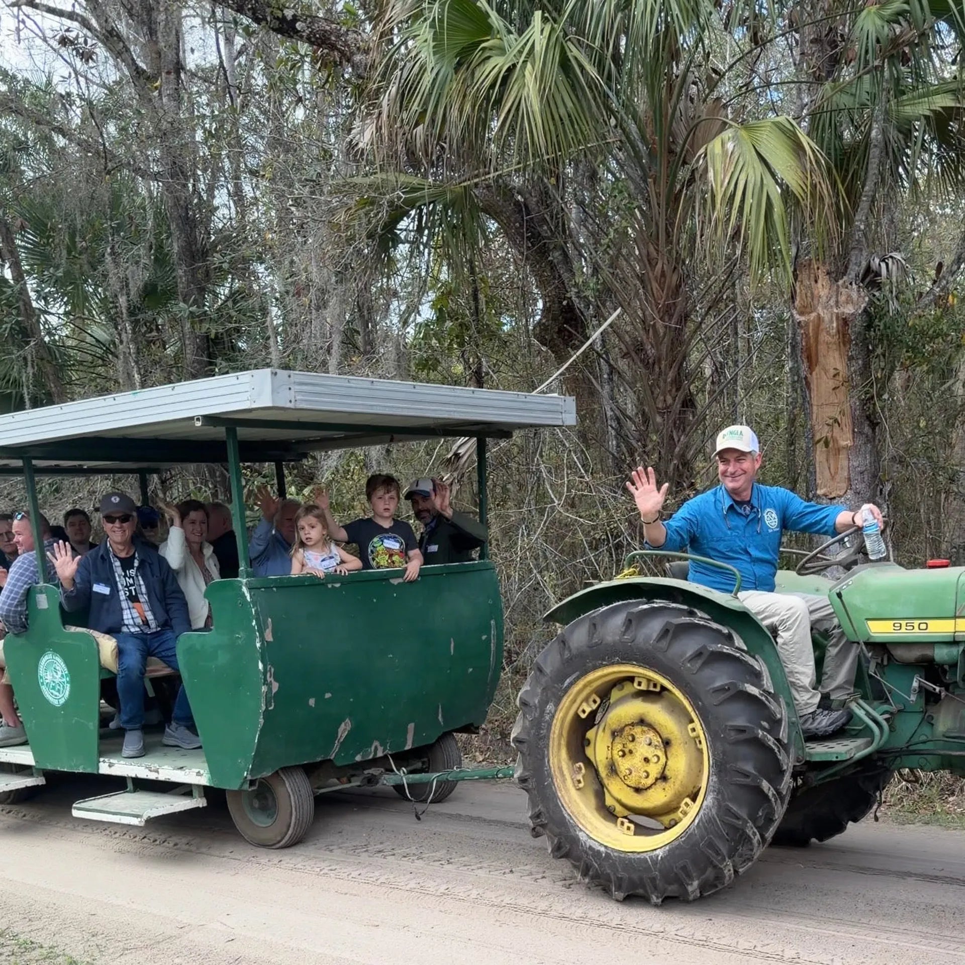 A green tractor towing a green passenger cart filled with people on a dirt road, with trees and palm foliage in the background. A man in a blue shirt and white cap sits in the tractor and waves.