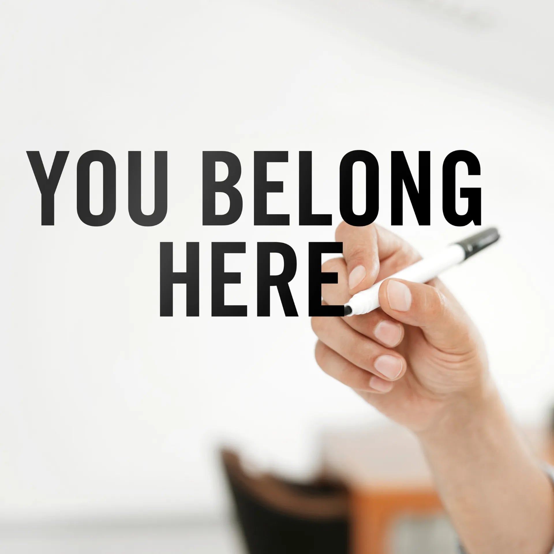 A hand holding a white marker writes the words 'YOU BELONG HERE' in large black capital letters on a whiteboard, with a softly blurred indoor background.