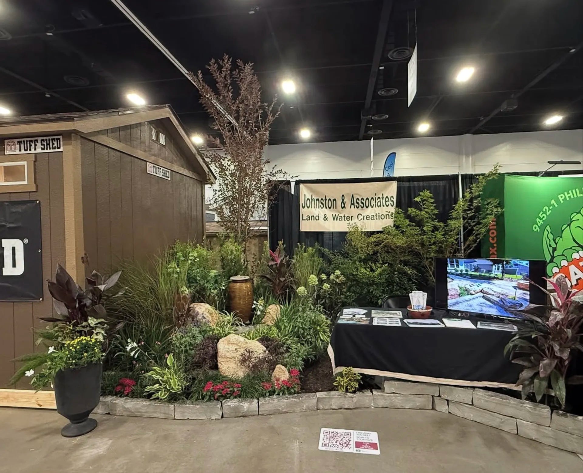 Exhibition booth with plants, a TV screen, and branded banners in an indoor setting.